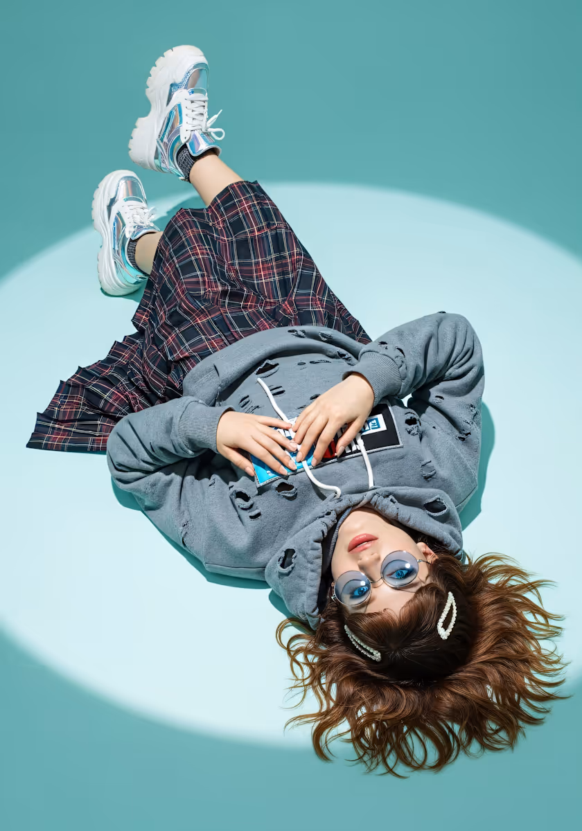 Young woman wearing blue tinted glasses, gray distressed hoodie, plaid skirt, and chunky sneakers lying on a mint green floor with a spotlight circle.